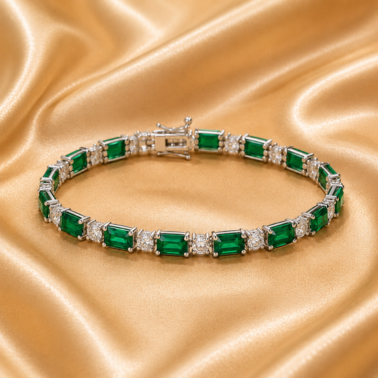 A silver tennis bracelet featuring emerald-cut green gemstones and small clear accent stones, arranged on a draped gold satin fabric background.