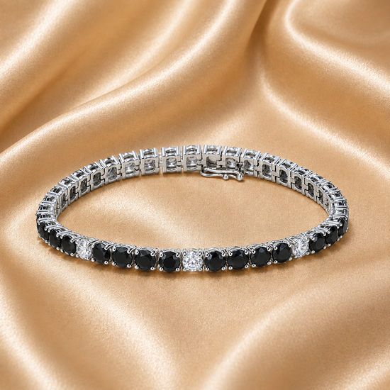 A tennis bracelet with alternating round black and clear gemstones in a silver-toned setting, displayed on a draped champagne satin fabric background.