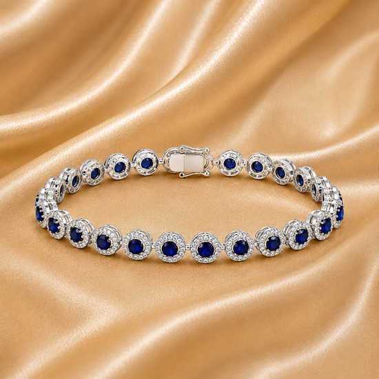A close-up of a silver tennis bracelet featuring round deep blue gemstones surrounded by clear crystal halo settings, displayed on a draped champagne satin fabric.