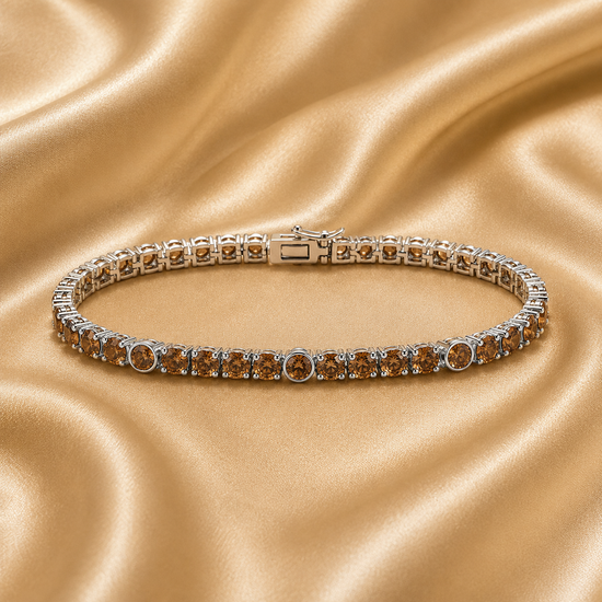 A silver tennis bracelet with round amber-brown gemstones, displayed on a draped champagne satin fabric background.