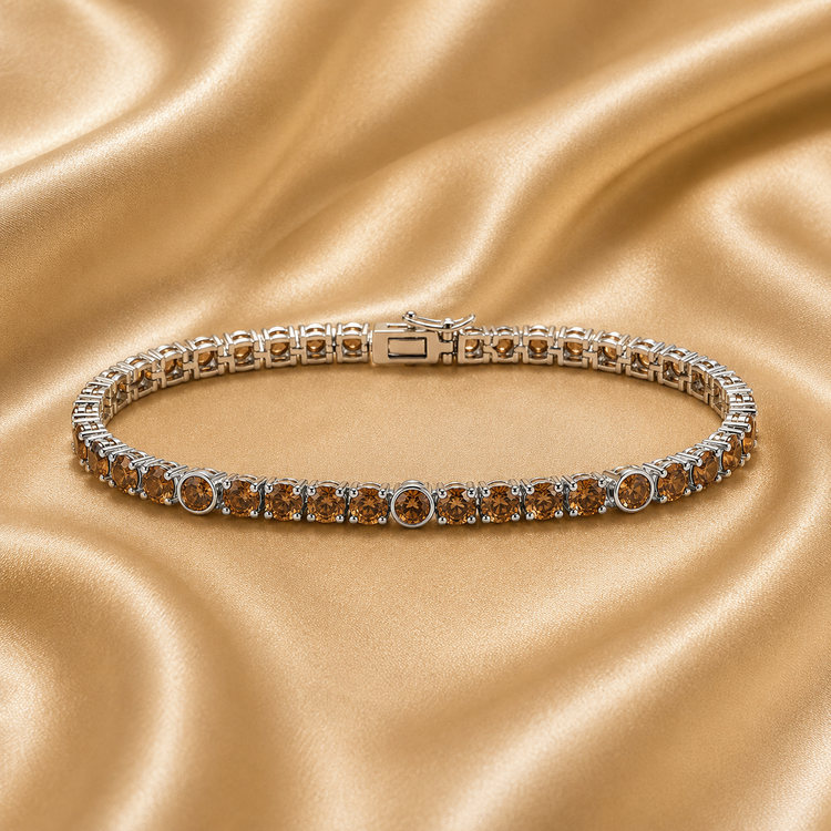 A silver tennis bracelet with round amber-brown gemstones, displayed on a draped champagne satin fabric background.