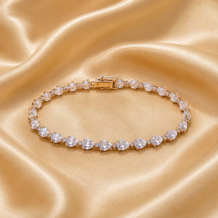 A gold tennis bracelet with clear round-cut gemstones, displayed on a draped champagne-colored satin fabric.