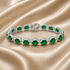 A silver tennis bracelet with oval green emerald-cut gemstones and small clear accent stones, displayed on draped beige satin fabric. The brand name &