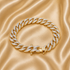 A gold-toned Cuban link chain bracelet paved with clear stones, displayed on a draped champagne satin fabric background.