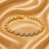 A gold chain bracelet with five pavé-set heart-shaped crystal clusters, displayed on a draped champagne-colored satin fabric.
