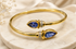Gold bangle bracelet with two blue teardrop gemstones and small clear accent stones, displayed on a light marble surface with soft fabric in the background.