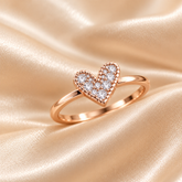 A close-up of a rose gold ring with a heart-shaped pavé setting worn on a finger, set against a soft beige satin fabric background.