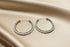A pair of textured silver hoop earrings displayed on a draped beige satin fabric background.