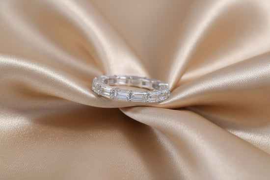 A silver band ring with baguette-cut clear stones, displayed on a draped champagne-colored satin fabric.