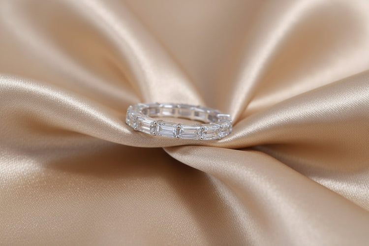 A silver band ring with baguette-cut clear stones, displayed on a draped champagne-colored satin fabric.