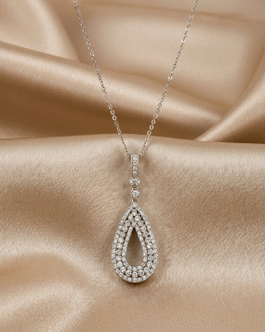 A silver teardrop pendant necklace paved with clear gemstones, resting on draped champagne-colored satin fabric.
