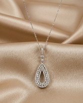 A silver teardrop pendant necklace paved with clear gemstones, resting on draped champagne-colored satin fabric.