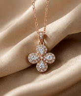 A rose gold four-petal flower pendant necklace encrusted with clear gemstones, resting on draped champagne-colored satin fabric.