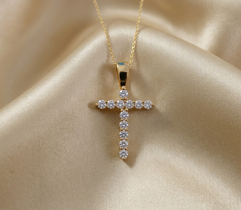 A gold cross pendant necklace adorned with clear gemstones, resting on a draped beige satin fabric background.