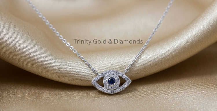 A silver-tone evil eye pendant necklace with a dark blue center stone and clear stones around the eye, resting on draped champagne-colored satin fabric. The text &