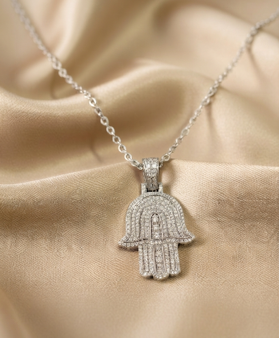 A silver Hamsa hand pendant necklace paved with small clear stones, displayed on draped champagne satin fabric.