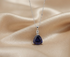 A silver pendant necklace featuring a deep teardrop-shaped dark blue gemstone and a small clear accent stone, displayed on draped champagne-colored satin fabric.