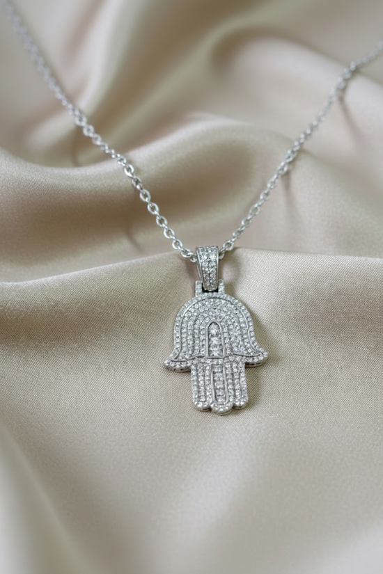 A silver Hamsa hand pendant necklace paved with clear stones, resting on draped champagne-colored satin fabric. The brand name &