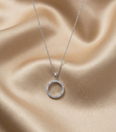 A silver chain necklace with a small open circular pendant set with clear stones, displayed on draped champagne satin fabric.