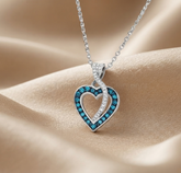 A silver heart-shaped pendant necklace with a vibrant blue gemstone border, resting on draped beige satin fabric.