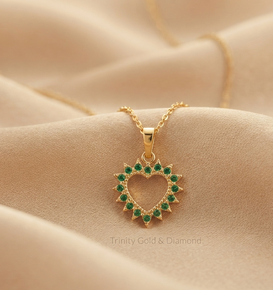 A gold heart-shaped pendant necklace with green gemstone accents, resting on a beige fabric surface. The brand name &
