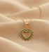 A gold heart-shaped pendant necklace with green gemstone accents, resting on a beige fabric surface. The brand name &