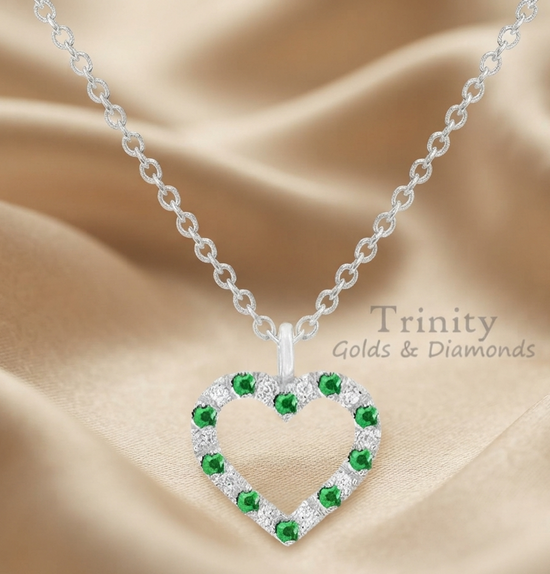 A silver heart-shaped pendant necklace with green and clear stones, displayed on a beige satin fabric background. The pendant features a silver heart outline with green gemstones on the left side and a border of clear stones on the bottom and right sides, leaving a hollow center.