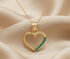 A gold heart-shaped pendant necklace with a curved row of green gemstones along the bottom edge, displayed on a soft beige satin fabric background. The brand name &